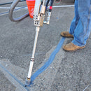 Close-up of the AST CMP 632 dispensing wand applying sealant to a concrete joint, with a worker in brown boots standing nearby and a hose connected to the system visible in the background.