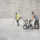 Two workers apply material using the AST GMP 050 dual-component pump system in a spacious warehouse. One person guides the application wand while the other manages the wheeled unit, which contains tanks for dispensing two-part epoxy, polyurea, or urethane.