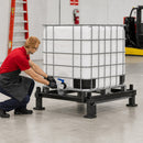 Worker dispensing liquid from an IBC tote positioned on a heavy-duty welded steel tote rack with integrated pump mounting area and 15-inch ground clearance in an industrial warehouse setting.